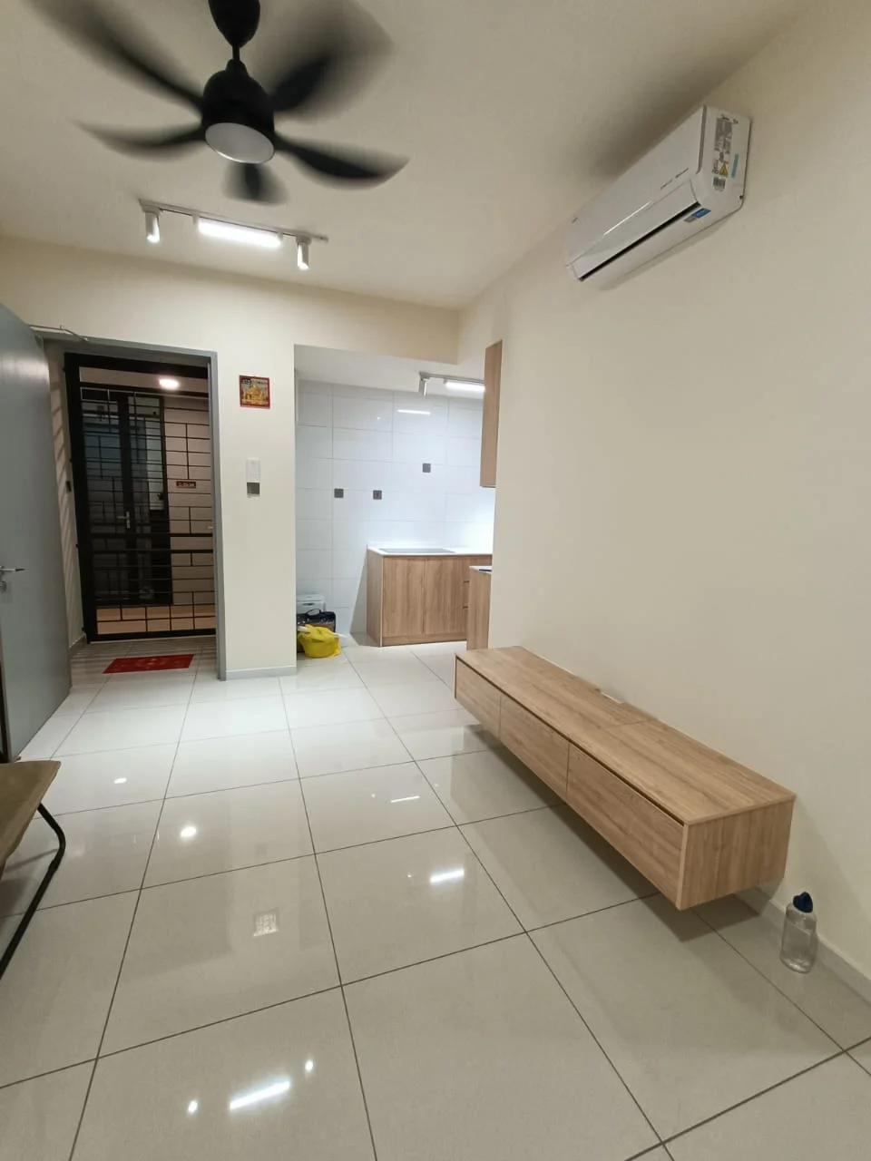 Room features: white painted walls, glossy ceramic tile flooring, 5-blade ceiling fan with light, wall-mounted split AC, wooden bench with storage, recessed ceiling lights, open layout, kitchen area visible in background; style: modern.